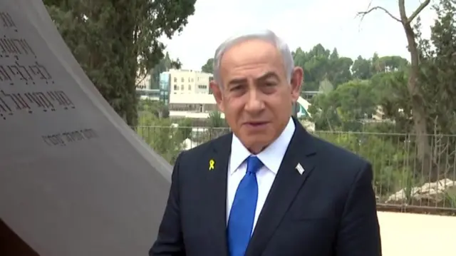 Benjamin Netanyahu mid-shot wearing black suit with royal blue tie. He wears a Israeli flag pin on left lapel and yellow hostage ribbon on right lapel