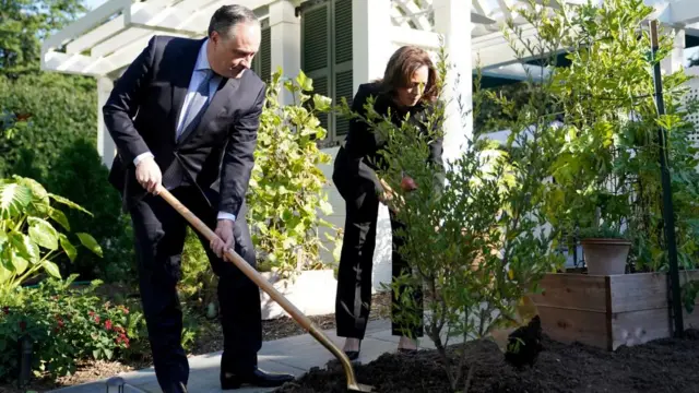 Douglas Emhoff and Kamala Harris both hold shovels and pick up dirt to put it on a tree in a garden