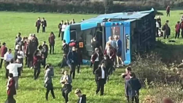 An image of a blue double decker bus lying on it's side in a field. There are a number of children surrounding it.