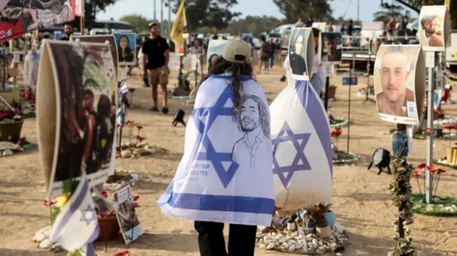 A woman walks while wearing a cream baseball cap and draped in an Israeli flag, as people visit the site of the Nova festival