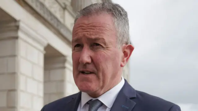 Murphy is wearing a nay suit with a white shirt and a blue and white tie. He's standing outside parliament buildings.