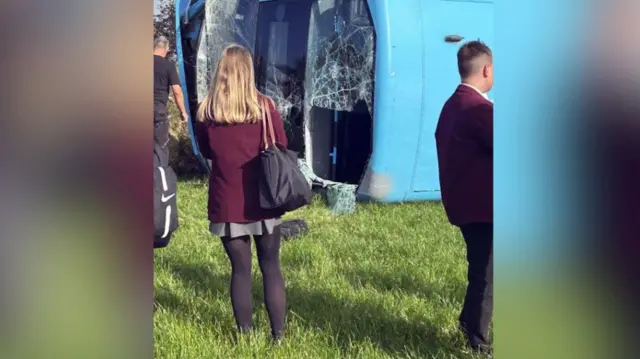 Two pupils stand in front of an overturned bus in a field