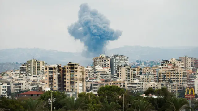 Smoke rises over some tower blocks in Tyre, with a McDonalds sign seen in the far right