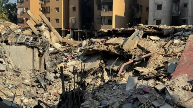 A picture of a street in Beirut, Lebanon, that has been bombed. Buildings lie ruined, there is shrapnel and rubble everywhere including a ruined car