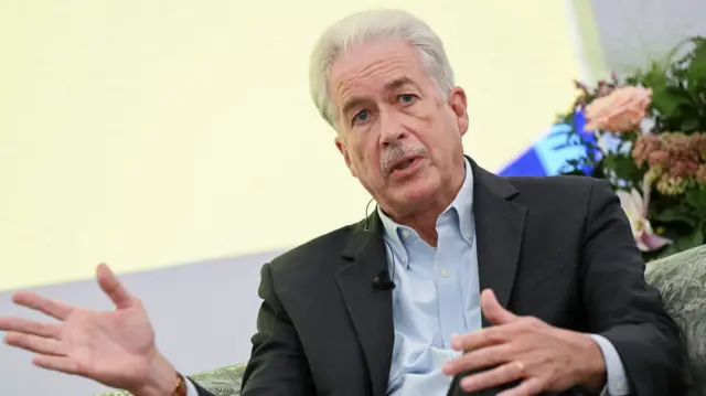 William Burns gestures with his hands as he speaks. He's sitting on a sofa with flowers behind him, and is wearing a blue shirt and black suit jacket. He has a moustache and white hair