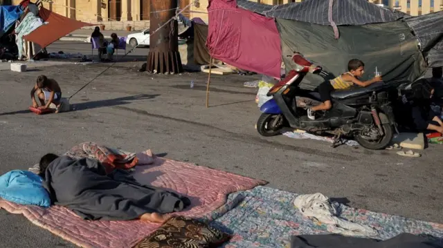 one child sleeps on a pink duvet laid on the ground while another sits on top of a moped while holding a plastic cup