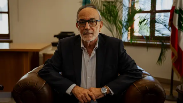 A man wears a suit jacket and shirt and sits in a leather armchair in an office, with a pot plant in the background