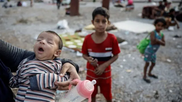 a woman holds a yawning infant, two other children blurred in background stare at the camera