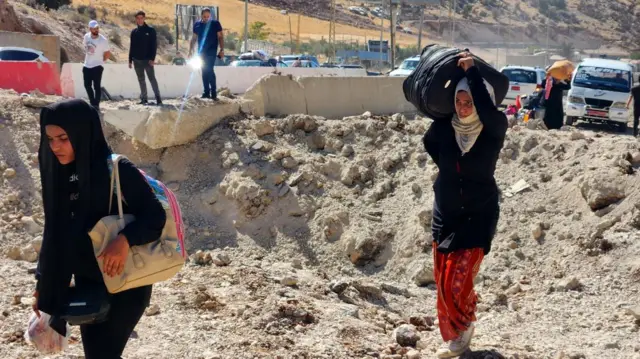 Displaced people carrying their belongings walk among debris near a shell crater at the Lebanon-Syria border