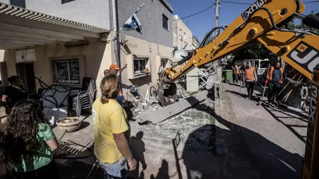 A person wearing a yellow t shirt watches as a digger helps to remove debris from a damaged house in Haifa