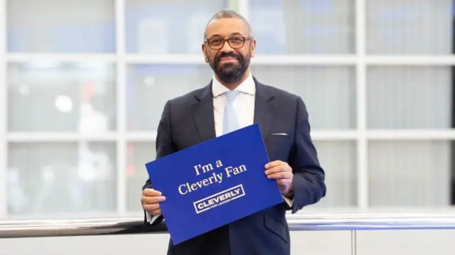 James Cleverly holding campaign merchandise, a sign that says "I'm a Cleverly Fan"