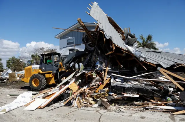 Yellow bulldozer pushes pile of debris together in front of light blue home in Keaton Beach