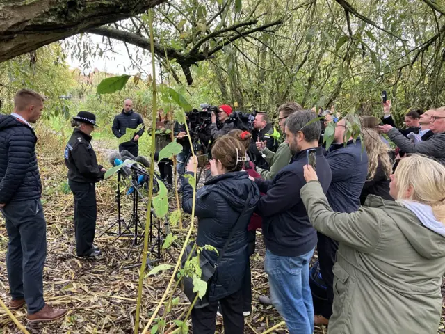 Reporters using cameras and phone gather around a police officer in front of a microphone in a wooded area