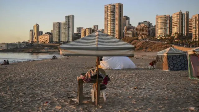 Tents and an umbrella are erected on a beach to shelter displaced Lebanese people