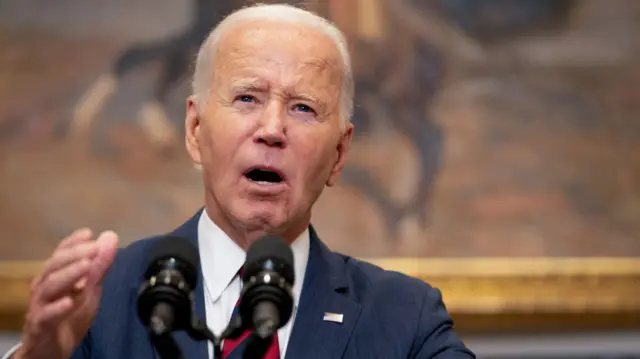 Bide, wearing a blue suit and red and blue striped tie, gestures as he addresses reporters in the White House