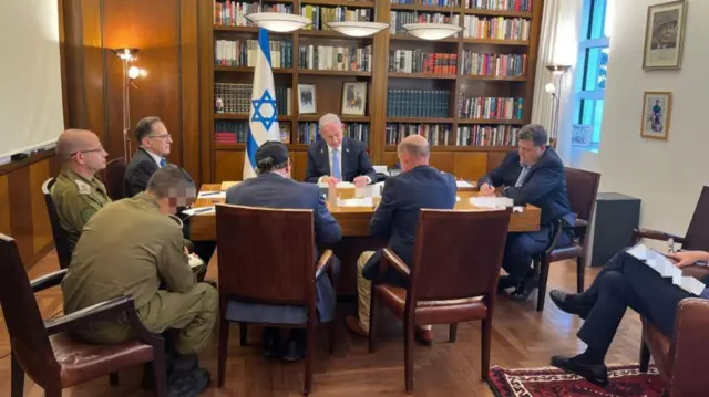 Netanyahu sits at a desk, surrounded by other men in suits, with a bookcase and Israeli flag in the background