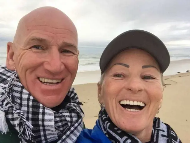 Leonard Nugent with his wife taking a selfie on a beach. They're both wearing black and white scarves around their necks