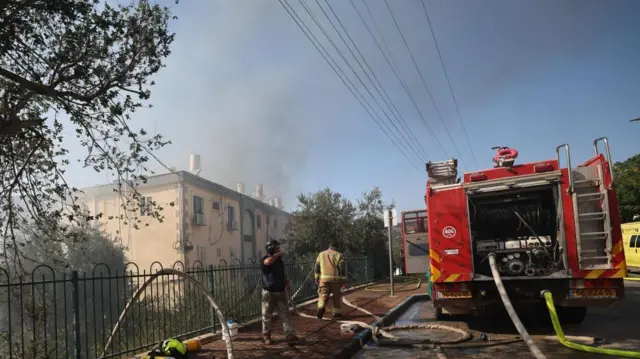 Firefighters work to put out a blaze in a residential building. They are standing on the street with fire hoses and a red fire truck