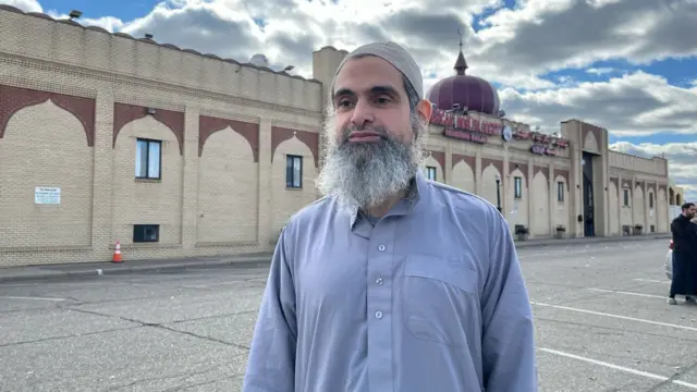 Hassan Abdel Salam, in traditional Muslim garb with a long grey beard, stands in front of a mosque