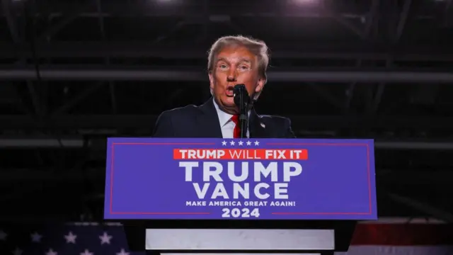 Donald Trump stands in front of a podium that reads: Trump/Vance Make American Great Again 2024