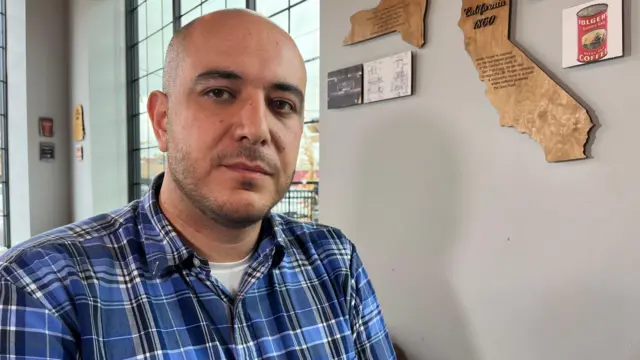 Abbas Alawieh wearing a blue checked shirt, sitting in front of a grey wall