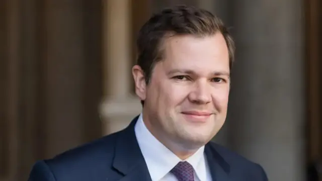 Robert Jenrick smiling while wearing a navy suit and purple tie