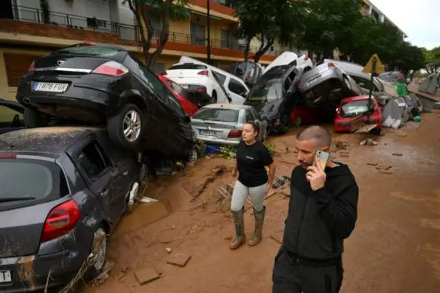 People walk past piles of cars swept up after the recent flash flooding in the nearby municipality Alfafar