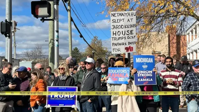 A crowd holding various anti-Trump and pro-Harris signs gathers behind police caution tape