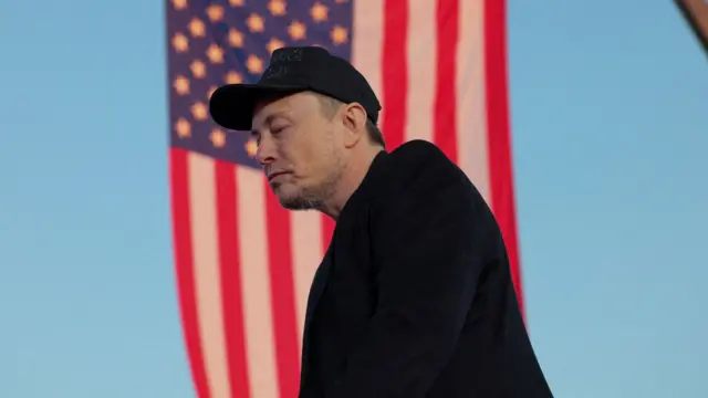 Elon Musk wearing black cap and jacket walking forward with USA flag waving behind him in the background