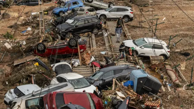 Cars littered across a railway line, some upside down, with other debris on the ground. People walk amongst the wreckage