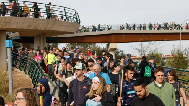 Large numbers of people crossing a bridge, some holding cleaning supplies and water