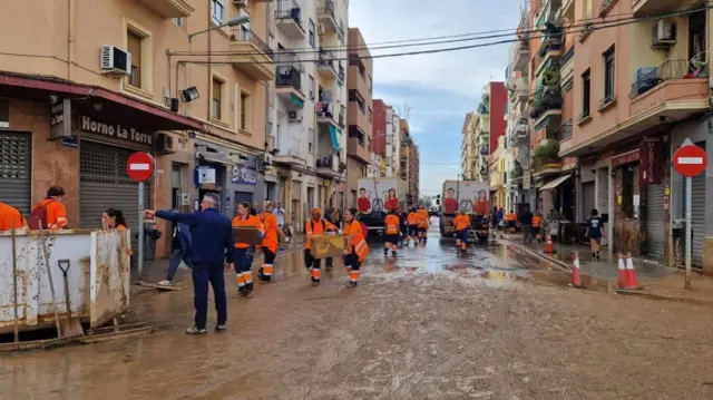 A muddy street in Valencia, there are workers in orange high vis wear arranging a cleanup of the street