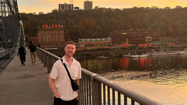 Newsbeat reporter Jordan Kenny standing on a bridge in Pittsburgh, Pennsylvania. The sun is setting in the background and people are walking across a bridge behind him.