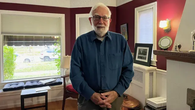 Prof Ron Stockton, in glasses and a blue shirt, stands in a living room looking at the camera