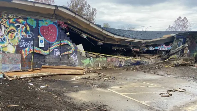 A building is seen with yellow police tape surrounding it and the roof is collapsing. There is graffiti on the outside of the building.