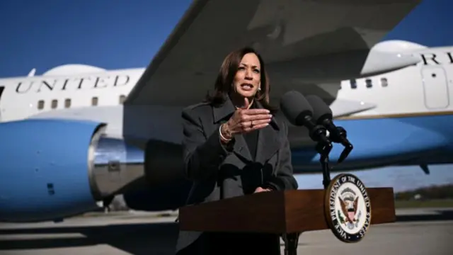 Harris speaking on a tarmac at a podium with the US presidential seal at the front, and Air Force 2 in the back.