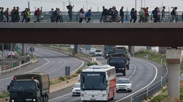 A group walks over a bridge as military vehicles pass underneath