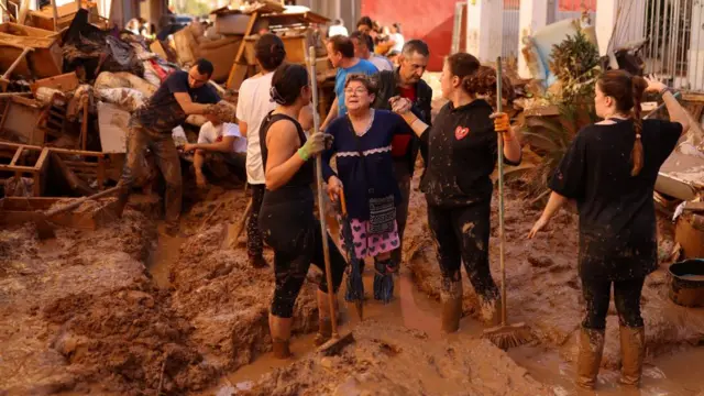 People stand in the street holding brooms, the mud is so thick it reaches their ankles