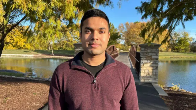 Dip Patel looks at the camera, with trees and a bridge in the background.