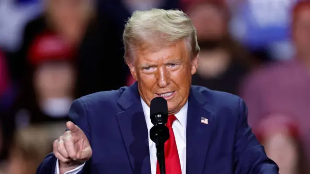 Donald Trump speaks on stage, his right hand extended and pointed finger pointing ahead. He's wearing a dark blue suit with white shirt and red tie. Behind him is a blurred group of supporters