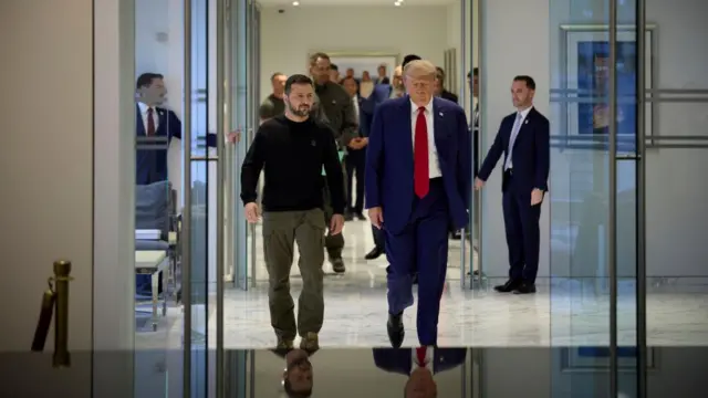 Zelensky and Trump walk side-by-side down a hallway. The Ukrainian president wears cargo pants and a black shirt, while Trump wears a blue suit and red tie.