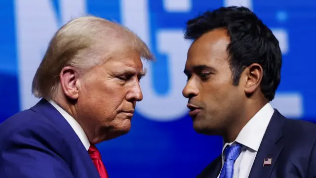 Donald Trump mid shot as he stands in front of Vivek Ramaswamy sideways. Both are seen from the top of the head to shortly below the shoulders. They're wearing dark blue suits