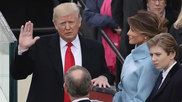 Donald Trump is sworn in as the new president in 2017. He's holding his right hand up, palm open, while his left hand is on the Bible. A smiling Melania Trump in blue dress coat holds the book, her son Barron Trump to her left