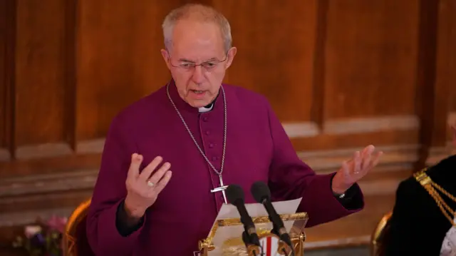 Justin Welby in purple robes and a cross around his neck gesture with his hands,