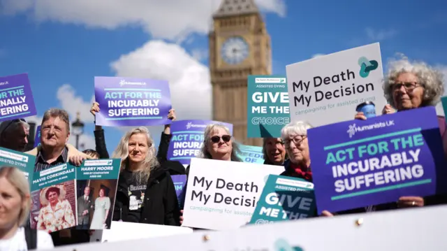 Assisted dying campaigners stand in a crowd with Big Ben in the background. They're holding placards that read 'my death, my decision' and 'act for the incurably suffering'