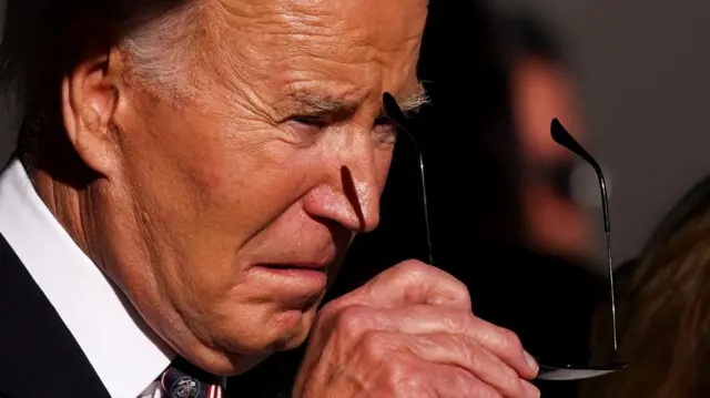 Joe Biden with the sun on his face holding his sunglasses in front of his head