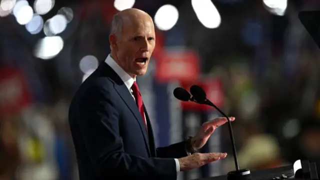 Side profile shot of Rick Scott talking at a lectern with his hands in front of him. He is wearing a dark suit and red tie. There are out of focus light behind him.