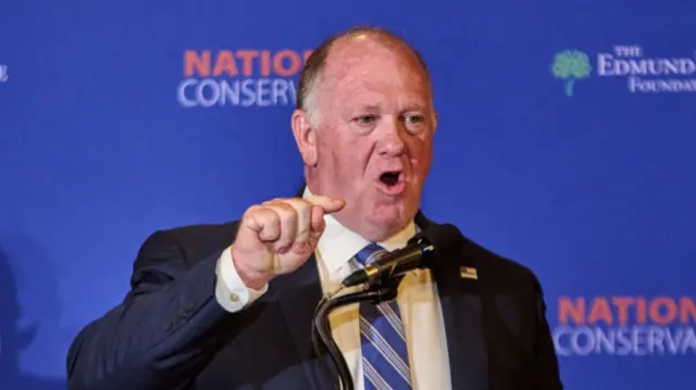 Tom Homan stood in front of a blue background, speaking into a microphone. He is pointing with his right hand as he speaks and is wearing a blue striped tie