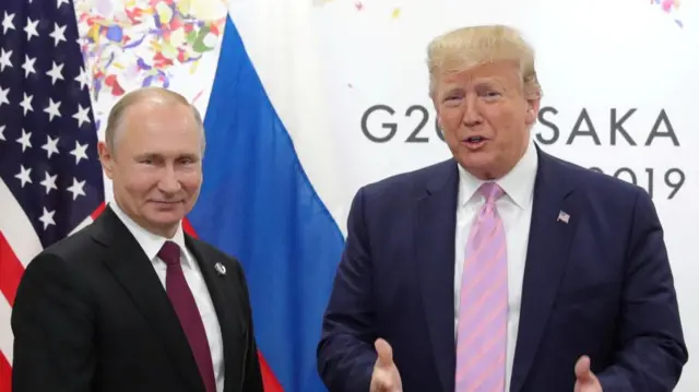 Vladimir Putin stands next to Donald Trump. Behind Putin, who is on the left hand side of the frame, are American and Russian flags. Trump, wearing a pink tie, stands in front of summit signage.