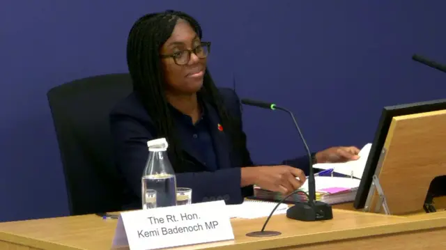 Kemi Badenoch sat at the inquiry witness desk, holding paperwork and a pen